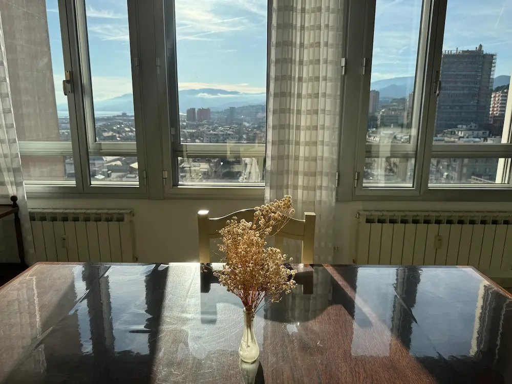 Un vaso con fiori secchi su un tavolo riflettente, con grandi finestre che rivelano un paesaggio urbano e le montagne sullo sfondo, che ricorda un elegante appartamento in affitto a Genova.